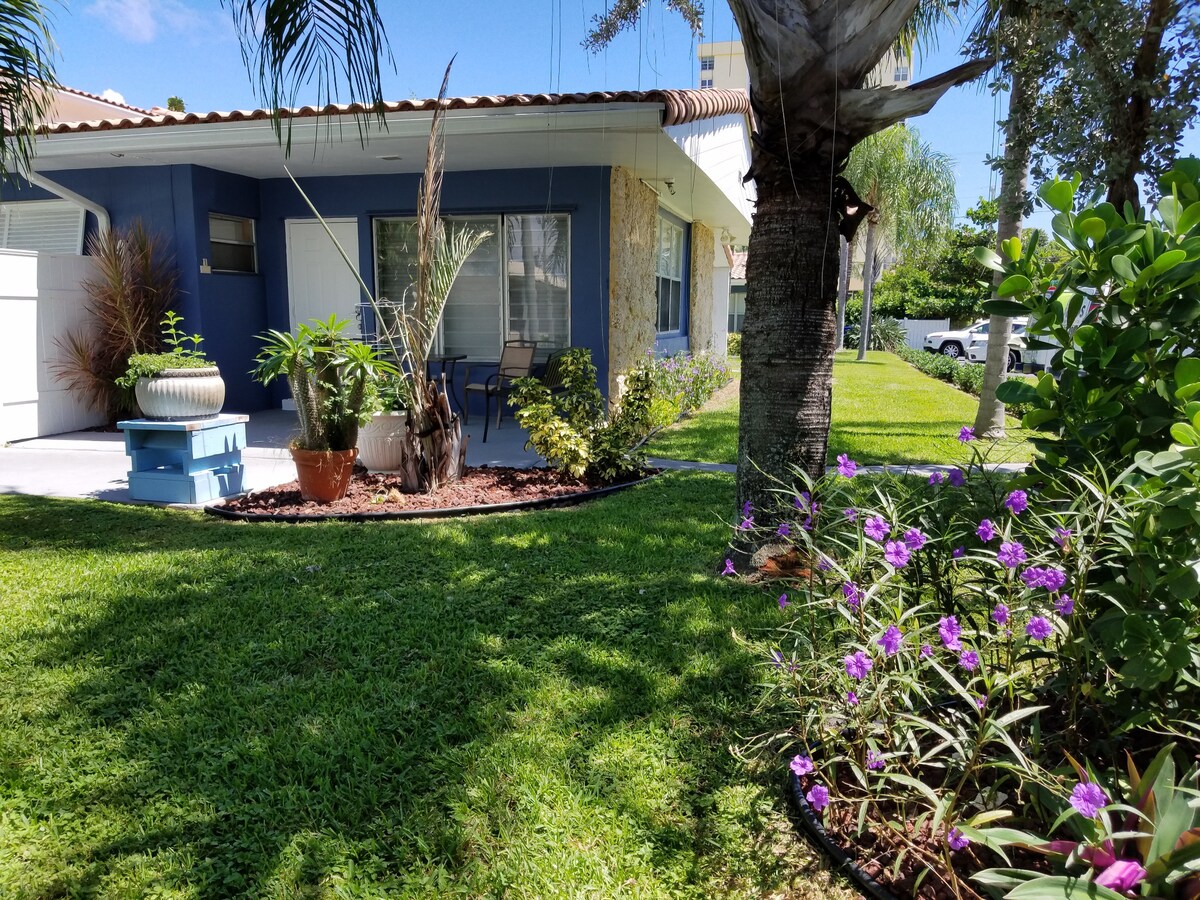 The exterior of the property features a well-maintained lawn with vibrant green grass. Decorative plants and flowers, including purple blooms, add color to the space. The building's blue exterior contrasts with the natural surroundings, creating a welcoming and serene atmosphere.