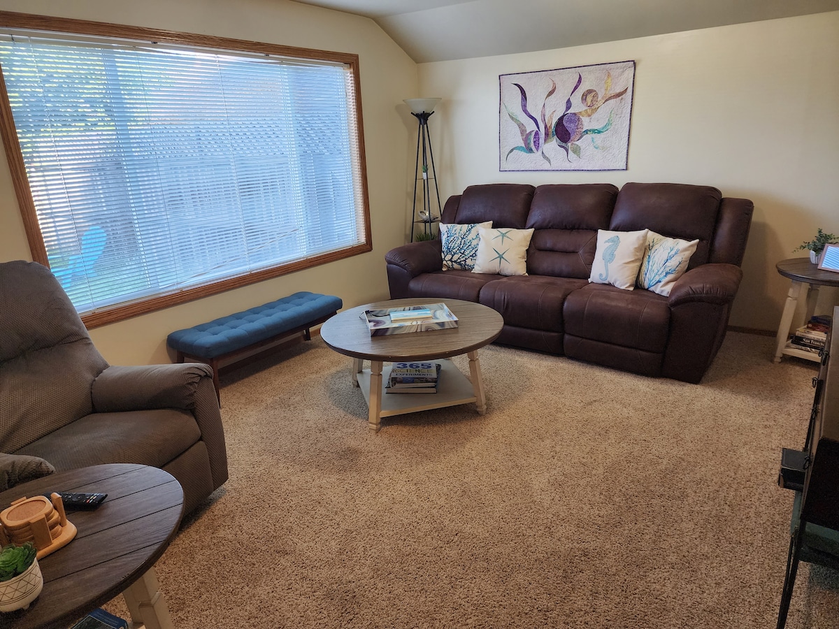A cozy living room is shown, featuring a brown sofa with decorative pillows, a round coffee table, and a blue bench. Natural light enters through a large window with blinds. A comfortable armchair and a lamp complete the inviting atmosphere.