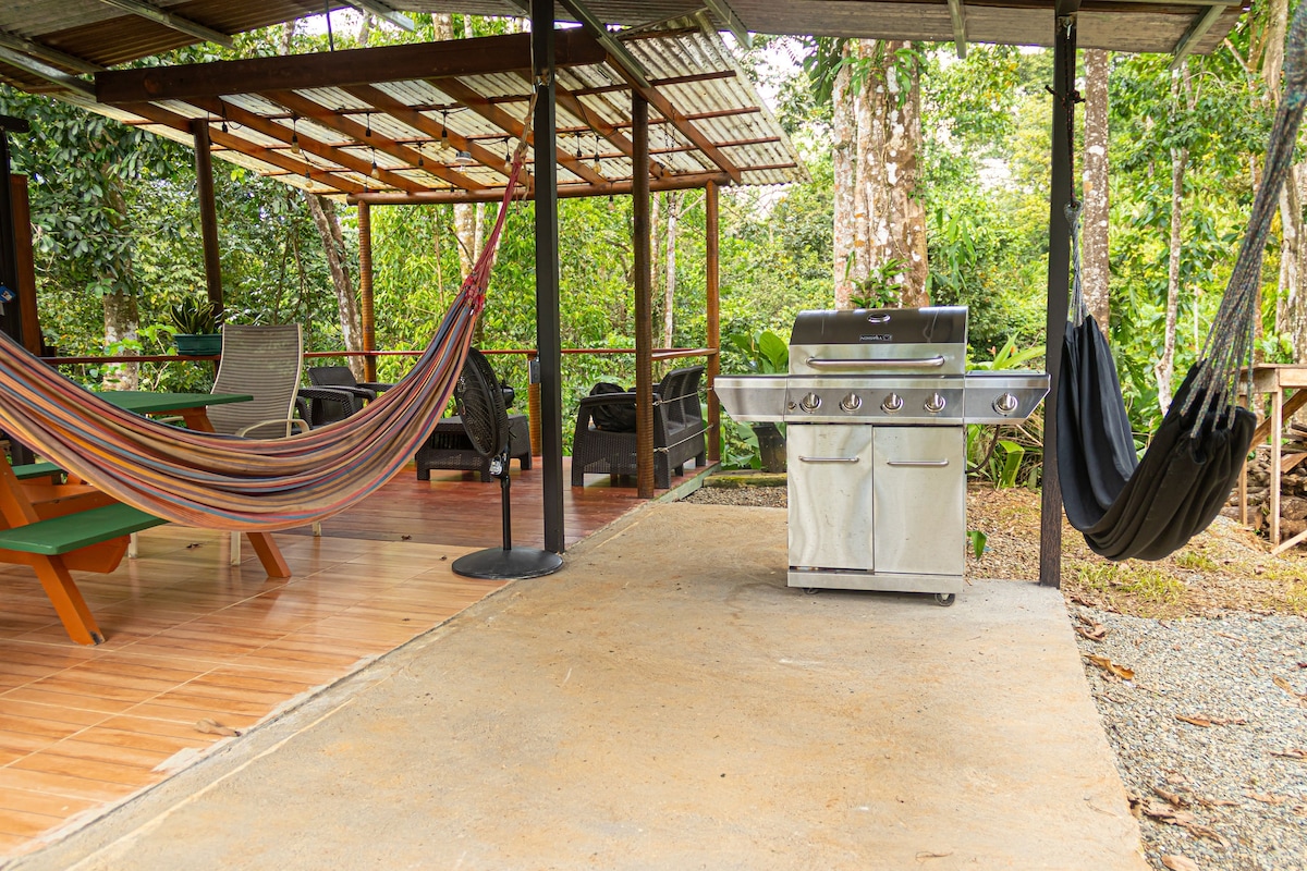 An outdoor space features several hammocks, a barbecue grill, and seating arrangements under a shaded structure. Lush greenery surrounds the area, creating a peaceful environment ideal for relaxation and outdoor cooking.