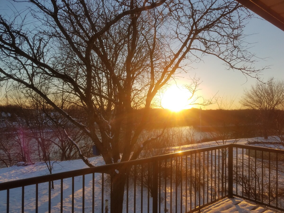 A tranquil sunset is viewed from a second-story balcony, casting golden light across the snowy landscape. Surrounding trees frame the scene, creating a serene atmosphere by the river's edge.
