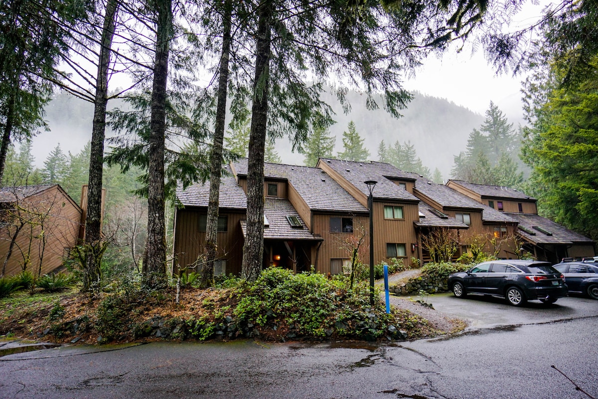 The exterior of the condo is surrounded by tall trees and greenery, reflecting a natural setting. A foggy mountain background enhances the serene atmosphere. Parking spaces are visible in front of the building, accentuated by a well-maintained driveway leading to the entrance.