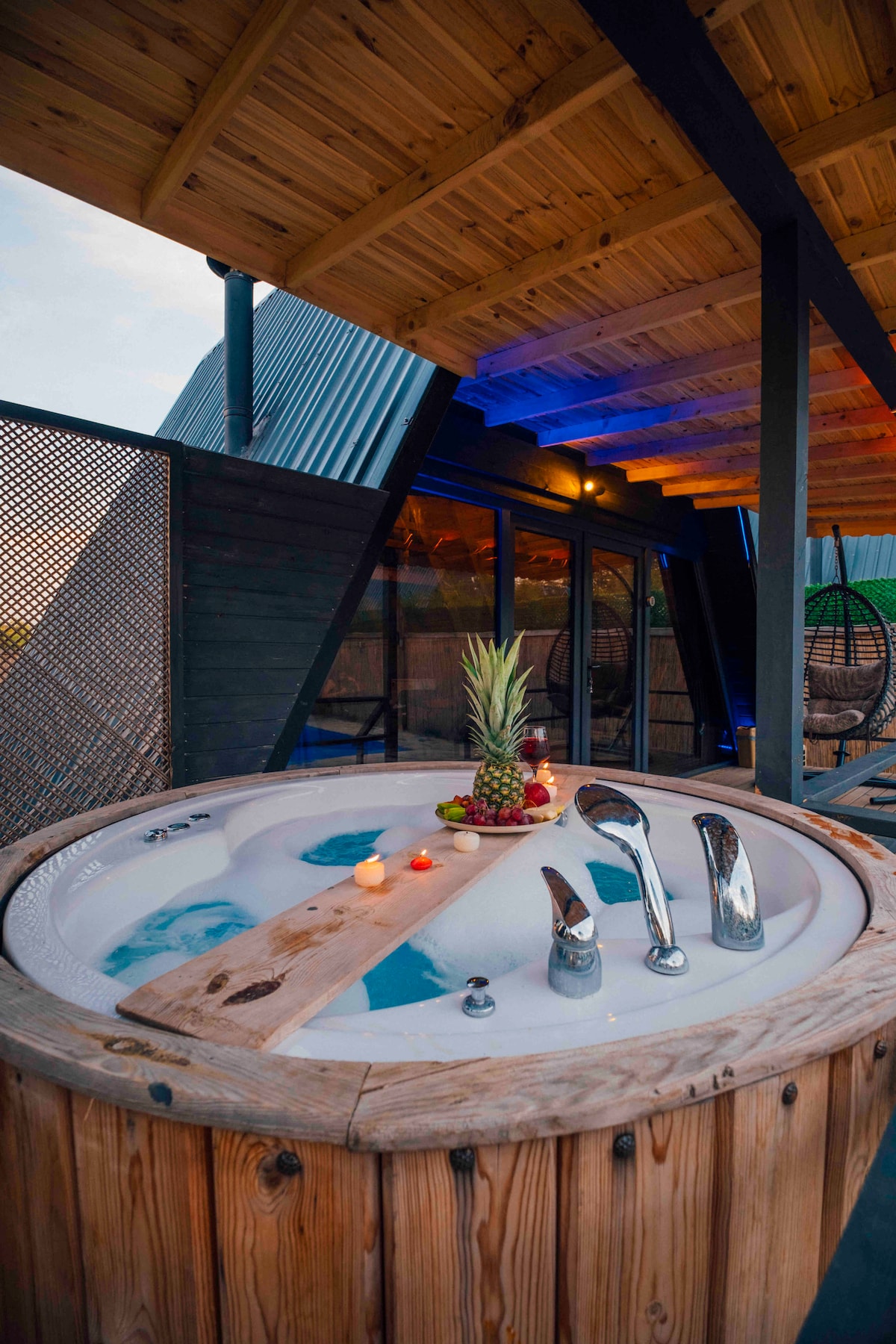 A wooden hot tub is surrounded by a wooden deck, featuring fruit and candles arranged on a wooden plank. Soft ambient lighting enhances the scene, while large glass doors provide views of the surrounding area.