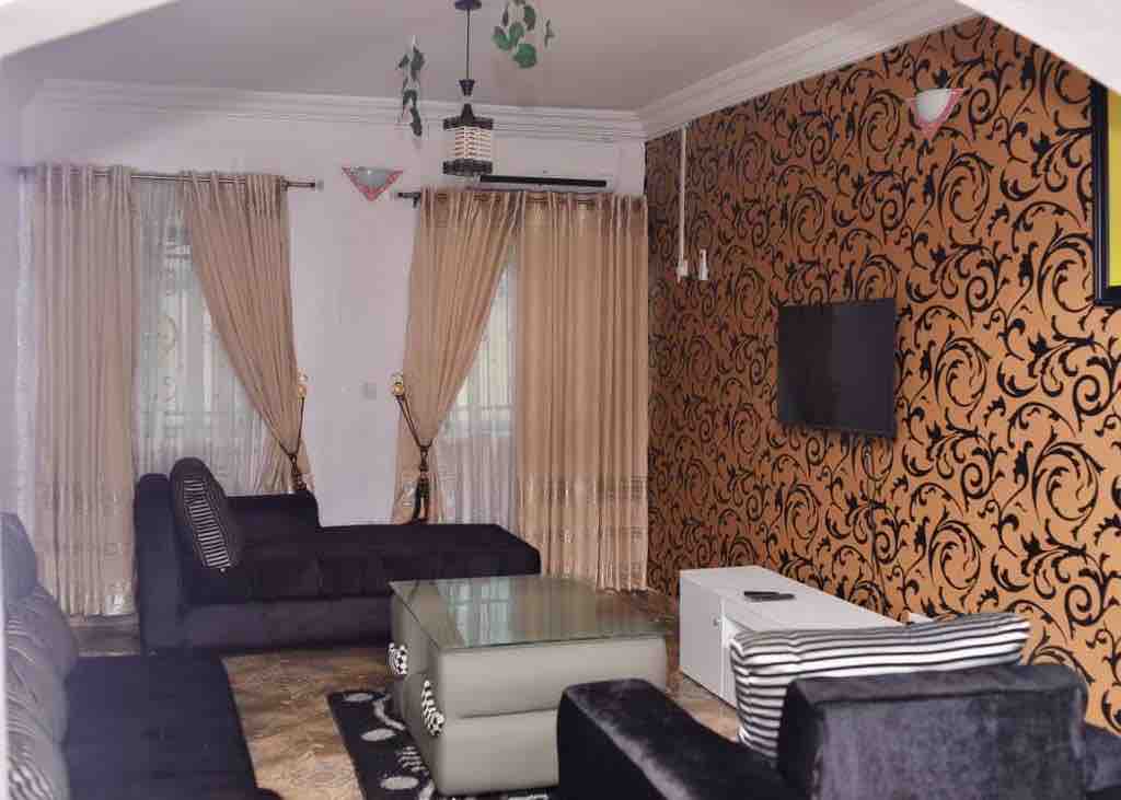 A spacious living area is adorned with decorative wallpaper featuring an intricate design. Two large windows are framed by sheer curtains, allowing natural light. A glass coffee table rests in the center, surrounded by several dark-colored couches, with a wall-mounted television for entertainment.