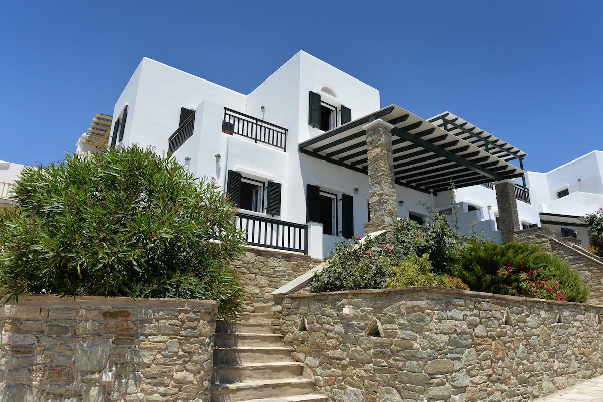 The two-story, Cycladic-style building features a whitewashed exterior with wooden balconies and green shutters. A stone wall surrounds the property, complemented by vibrant plants. A spacious terrace is shaded by a pergola, providing a welcoming entrance.