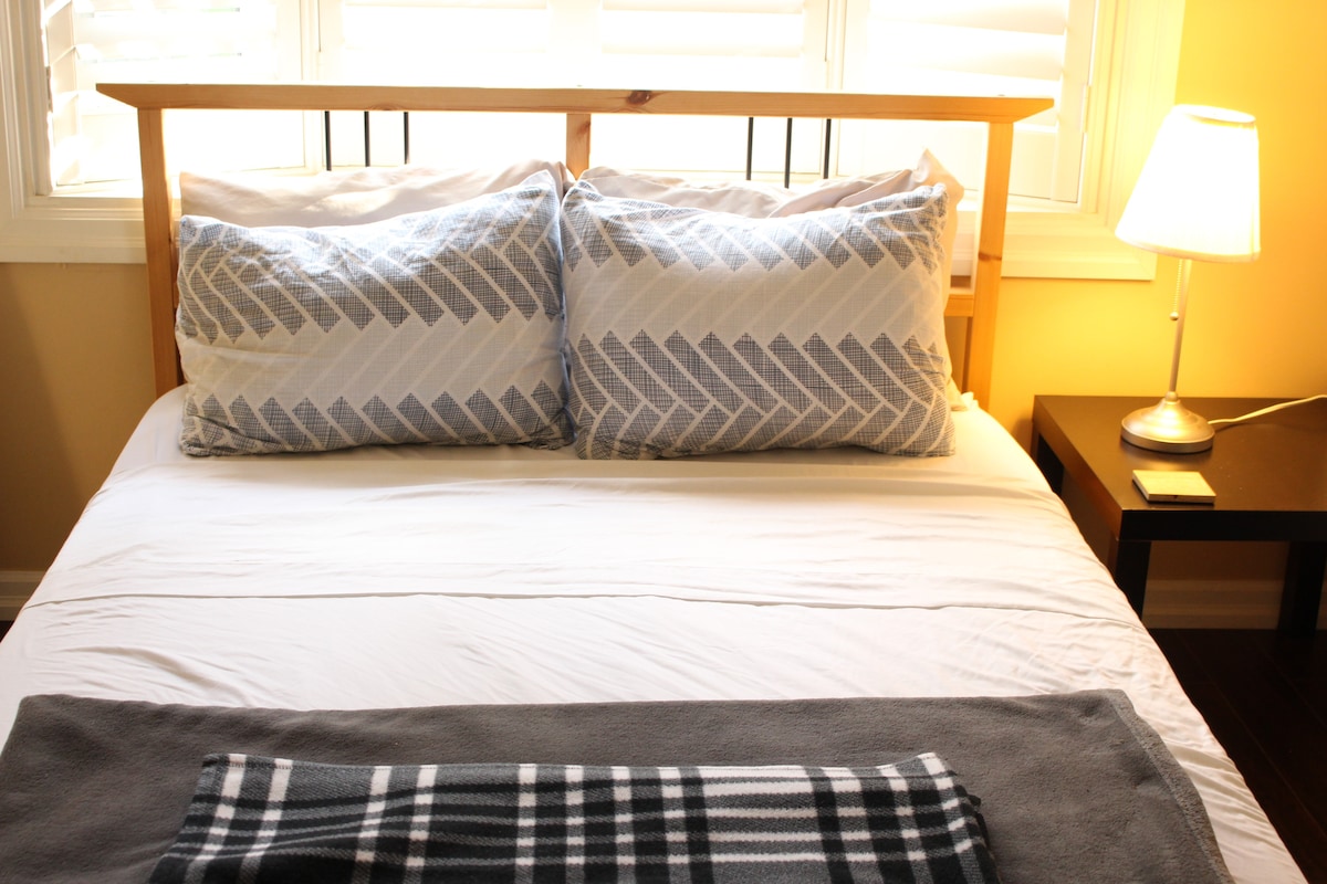 A comfortable bed is framed by soft pillows and a plaid blanket draped across the foot. A bedside lamp provides gentle illumination, while natural light filters in through partially closed window shutters, creating a serene atmosphere.