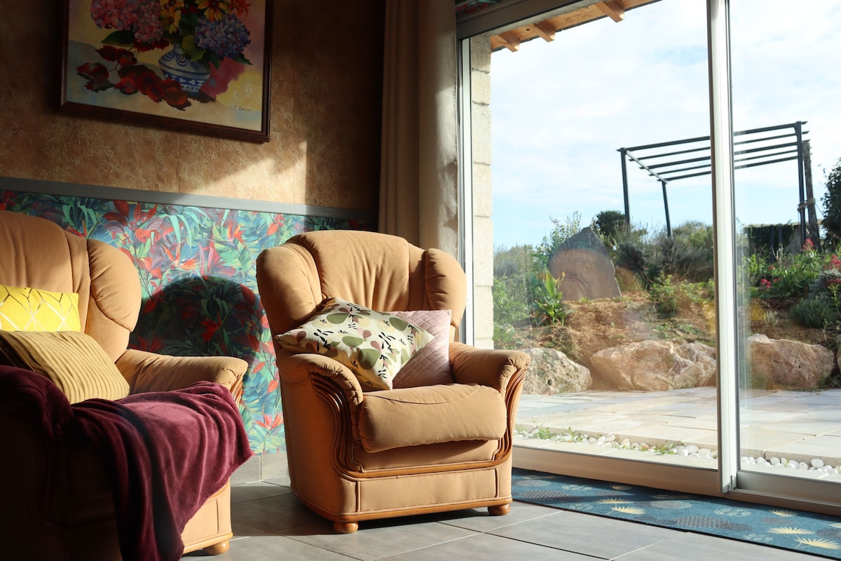 A cozy seating area is featured, showcasing two comfortable armchairs adorned with decorative cushions. Natural light filters through large glass doors, illuminating the room and providing a view of a landscaped outdoor area with greenery.