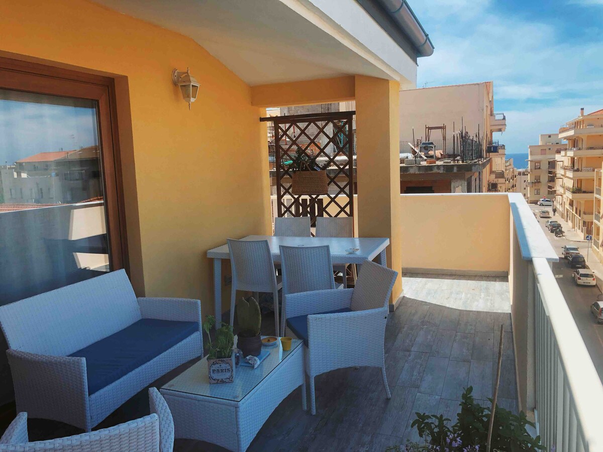A private terrace is presented with modern outdoor furniture, including a small table and seating area. The space is surrounded by a low railing, offering views of nearby buildings and a glimpse of the sea in the distance. Sunlight brightens the area.