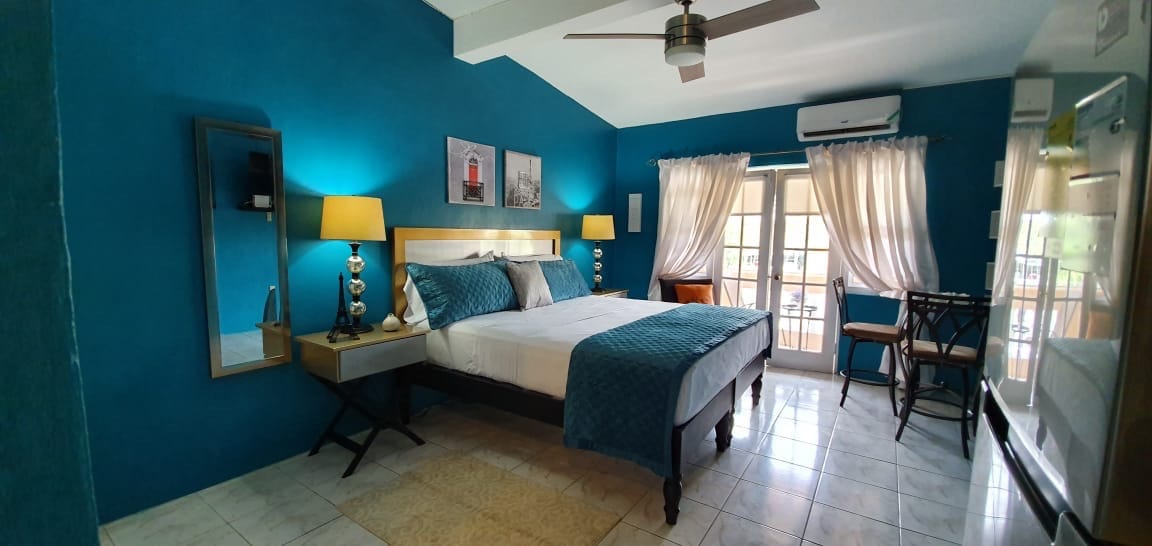 The studio features a spacious bed adorned with a textured blue throw and decorative pillows. A mirrored wall accent reflects warm light from two bedside lamps. Light-filtering curtains frame double glass doors that open to a private balcony, and a small dining table is positioned near the window.