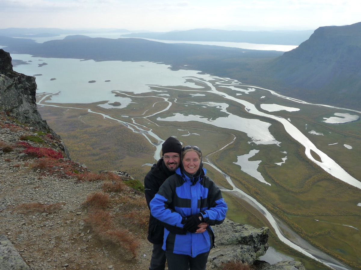 A breathtaking view from a mountain shows a winding river and a vast landscape stretching into the distance. Two individuals are positioned in the foreground, surrounded by rocky terrain and vibrant autumn colors, inviting a sense of adventure in a remote wilderness.