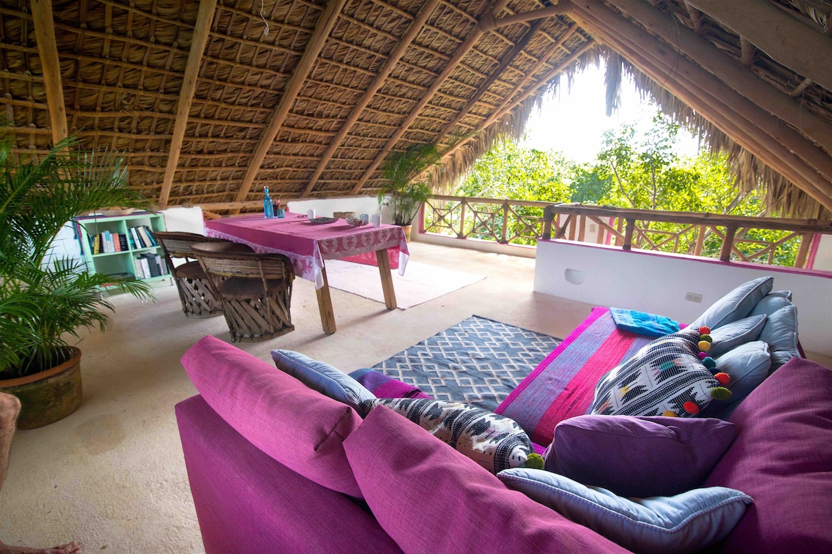 A spacious loft area features a thatched roof, offering cozy seating with colorful cushions and decorative pillows. A wooden table set for dining is visible, surrounded by natural greenery outside the large windows. Sunlight filters through, enhancing the inviting atmosphere.