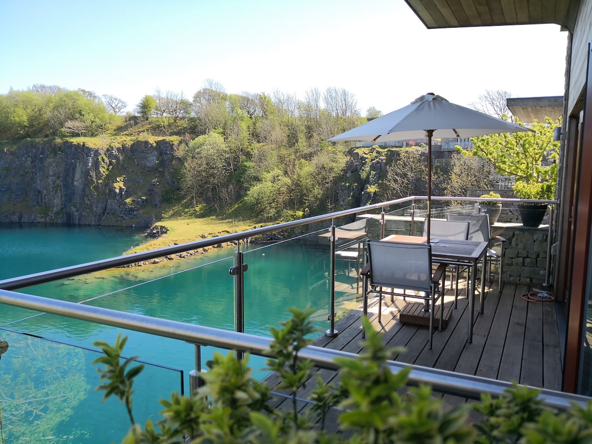 A wooden terrace features a dining set with an umbrella, overlooking a tranquil blue-green lake and lush greenery. The space is enclosed by glass railing, enhancing the view of the surrounding nature.