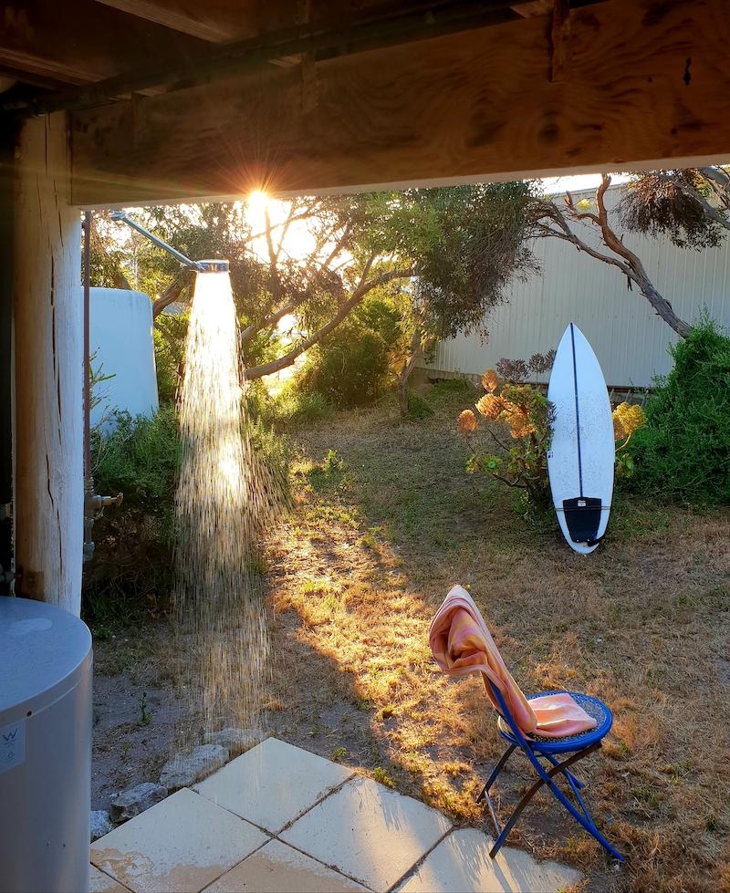 A spacious outdoor area features a shower with water cascading down, surrounded by greenery. A surfboard leans against a bush in the background. A chair sits nearby on a stone patio, offering a relaxing space to enjoy the natural setting.