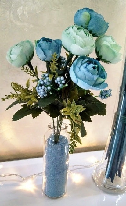 A decorative arrangement of artificial flowers is presented in a clear glass vase. The flowers feature various shades of blue and green, with leaves and berries complementing the design. Soft, ambient lighting glows in the background, enhancing the visual appeal of the arrangement.