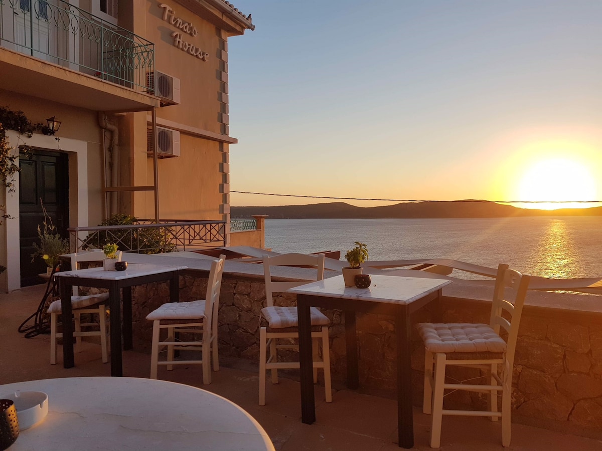 An outdoor seating area is depicted, featuring several white chairs and tables arranged against a backdrop of the setting sun. The tranquil sea is visible, reflecting warm hues, and potted plants add a touch of greenery to the space. The historic surroundings enhance the charm of the location.