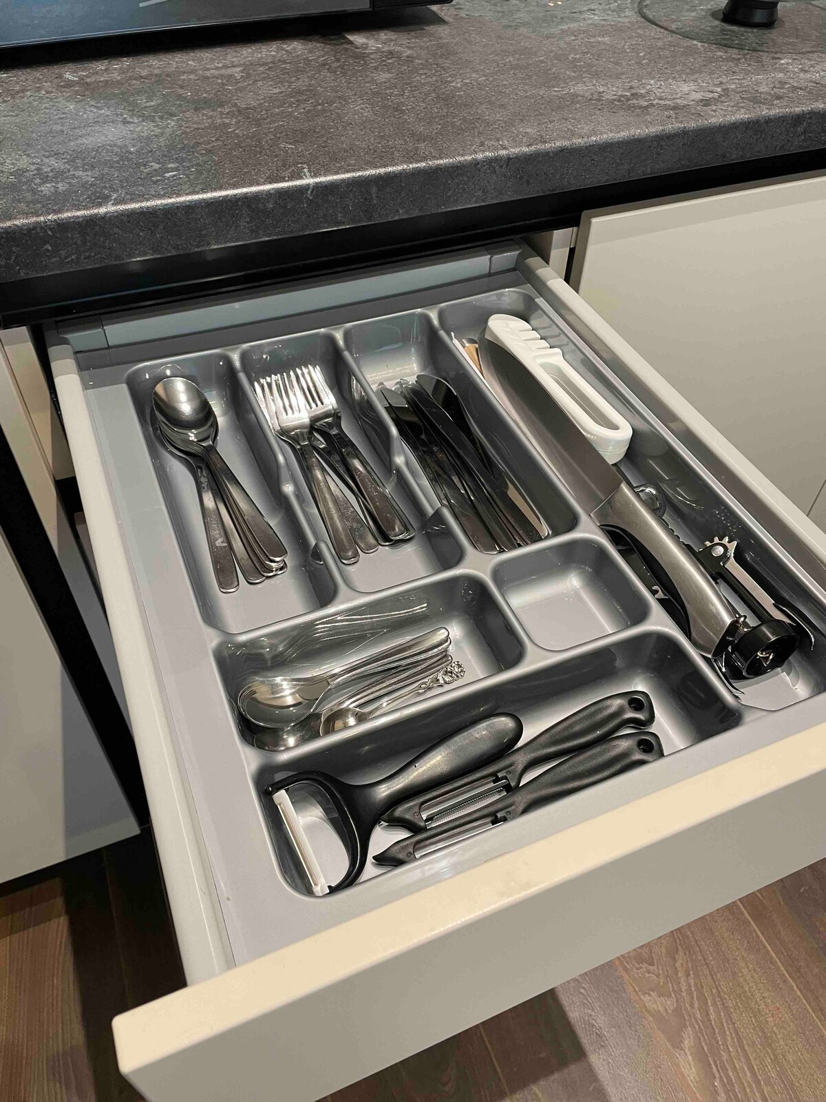 A kitchen drawer is shown open, containing an organized arrangement of cutlery and utensils. Different types of silverware, including forks, knives, and spoons, are neatly placed in designated compartments, along with additional cooking tools for convenience.