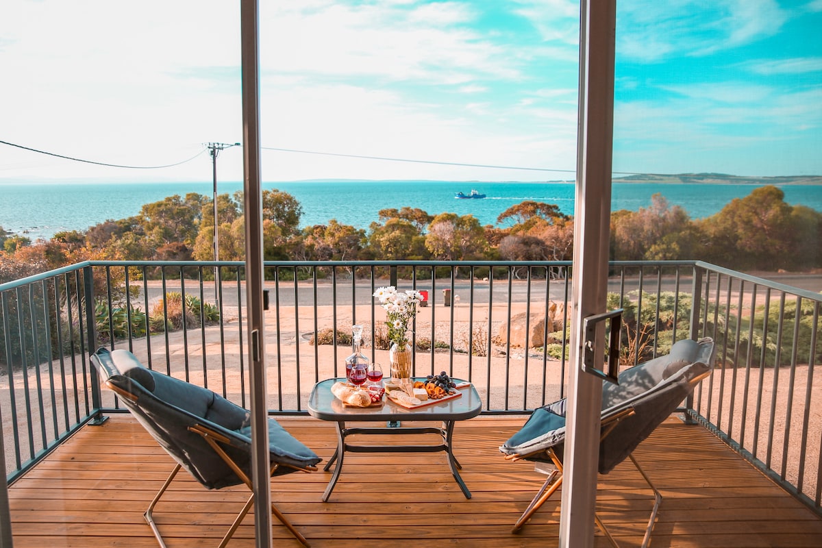 A balcony view showcases a table set for two, featuring a spread of snacks and drinks. Two chairs are positioned around the table, with the ocean visible in the background. Lush greenery and a glimpse of the coastline enhance the tranquil setting.