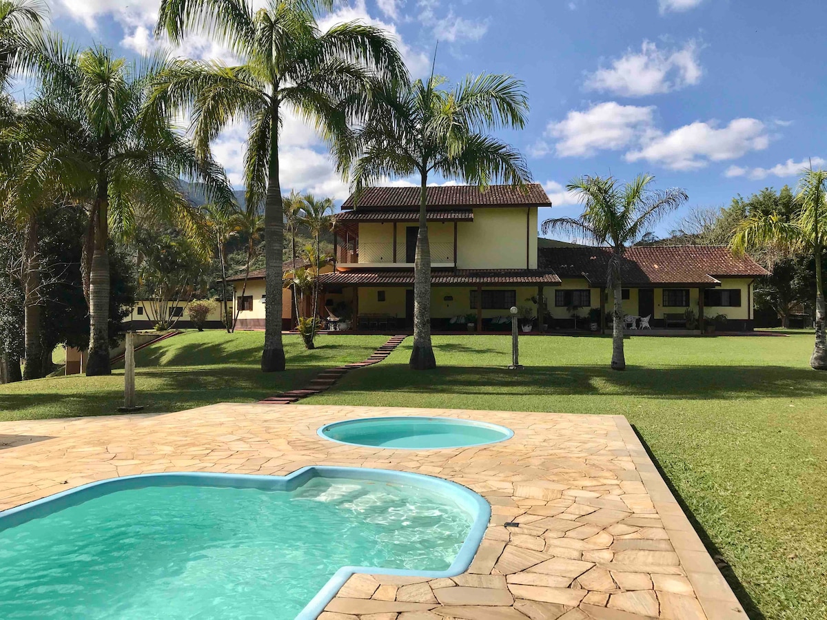 A view of the house surrounded by palm trees, featuring a spacious lawn. The outdoor area includes a distinctive pool with an adjoining smaller hot tub, set against a clear blue sky with scattered clouds.