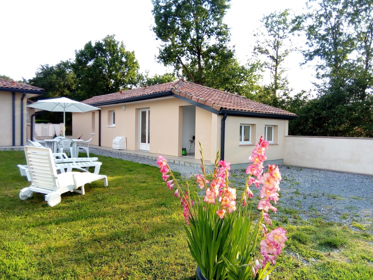 A quaint gîte is surrounded by lush greenery, featuring a bright exterior complemented by a tiled roof. A seating area with white lounge chairs is positioned on a gravel lawn, while colorful flowers in a planter add a touch of vibrancy to the scene.