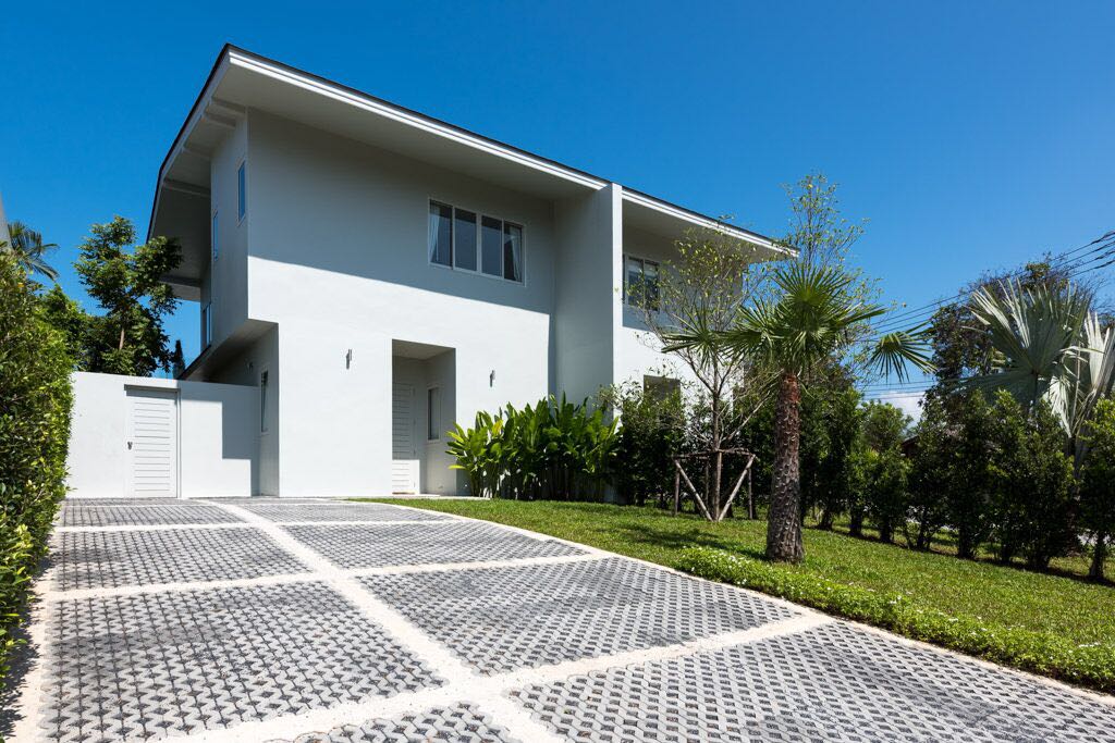 A modern two-story villa is depicted, surrounded by lush greenery and well-manicured landscaping. The driveway, composed of patterned pavers, leads to the entrance, with palm trees and bushes framing the property. Clear blue skies complete the inviting exterior.