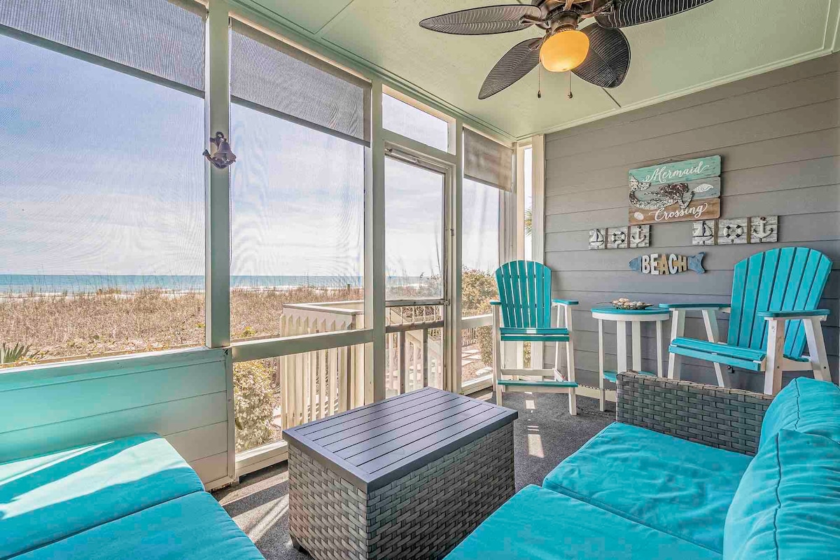 A spacious screened-in patio features turquoise chairs and a matching table. Sunlight streams through large windows, providing views of the beach and ocean. A neutral-colored rug adds warmth to the floor, while a modern ceiling fan enhances airflow.