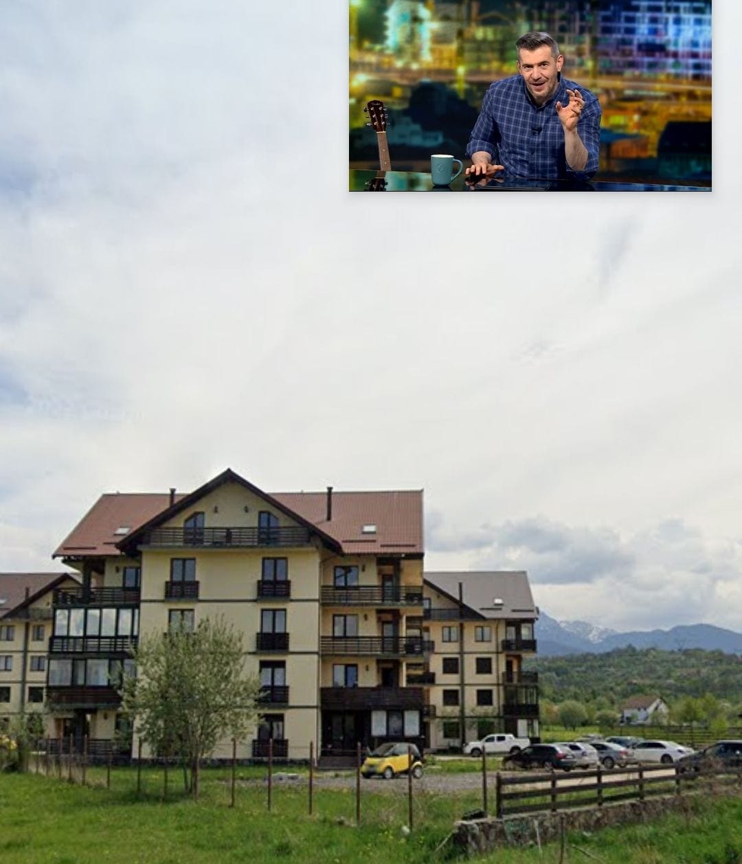 A modern multi-story building is set against a backdrop of rolling hills and cloudy skies. The building features a combination of balconies and large windows, offering scenic views. A grassy area in front is complemented by a small tree, while several parked cars are visible nearby.