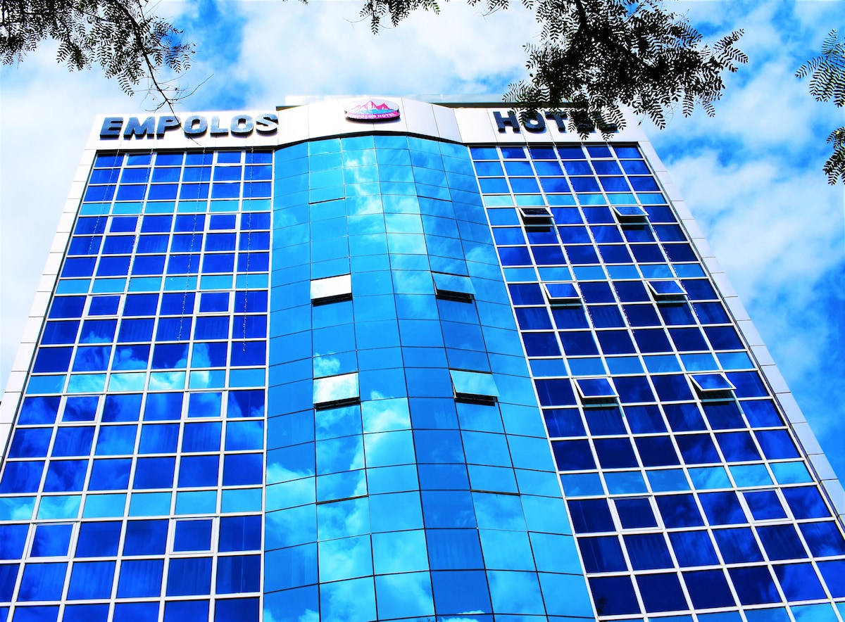 The exterior of Empolos Hotel is highlighted by a modern facade of reflective blue glass, capturing the sky and surrounding trees. The building features multiple stories, with prominent signage at the top that identifies the hotel.