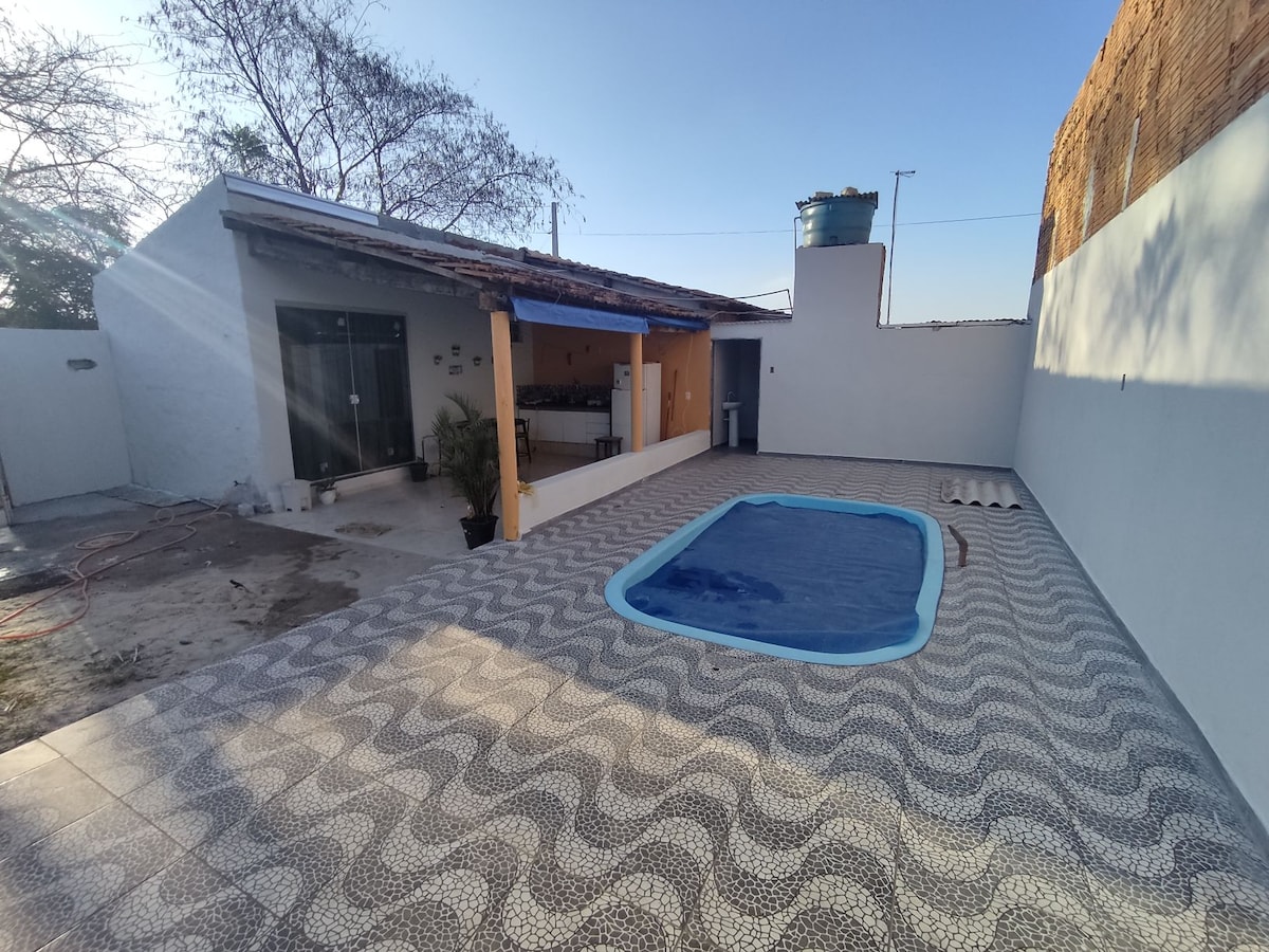 A serene outdoor area features a refreshing pool surrounded by patterned tiles. The sunlit space includes an entrance to an indoor area, with light filtering through glass doors. A water tank is positioned in the background against a clear blue sky.