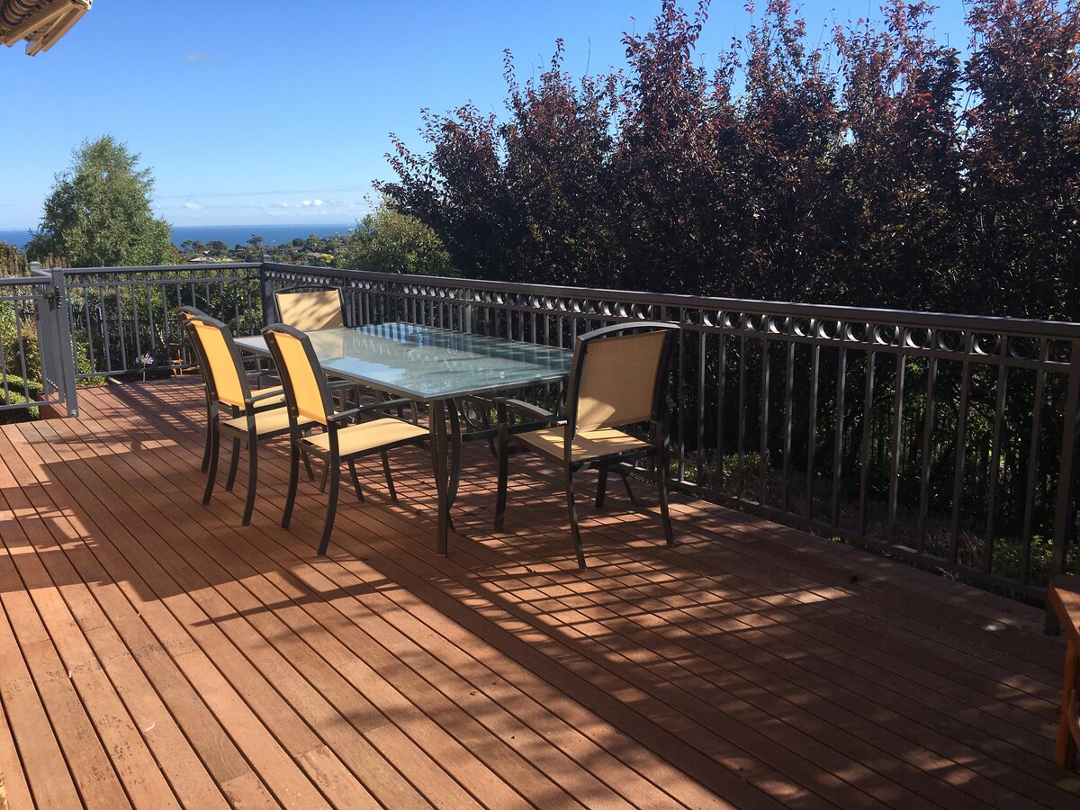 A spacious wooden deck is featured, designed for outdoor dining, with a glass-topped table surrounded by four chairs. Lush greenery and distant bay views provide a scenic backdrop under a clear blue sky.