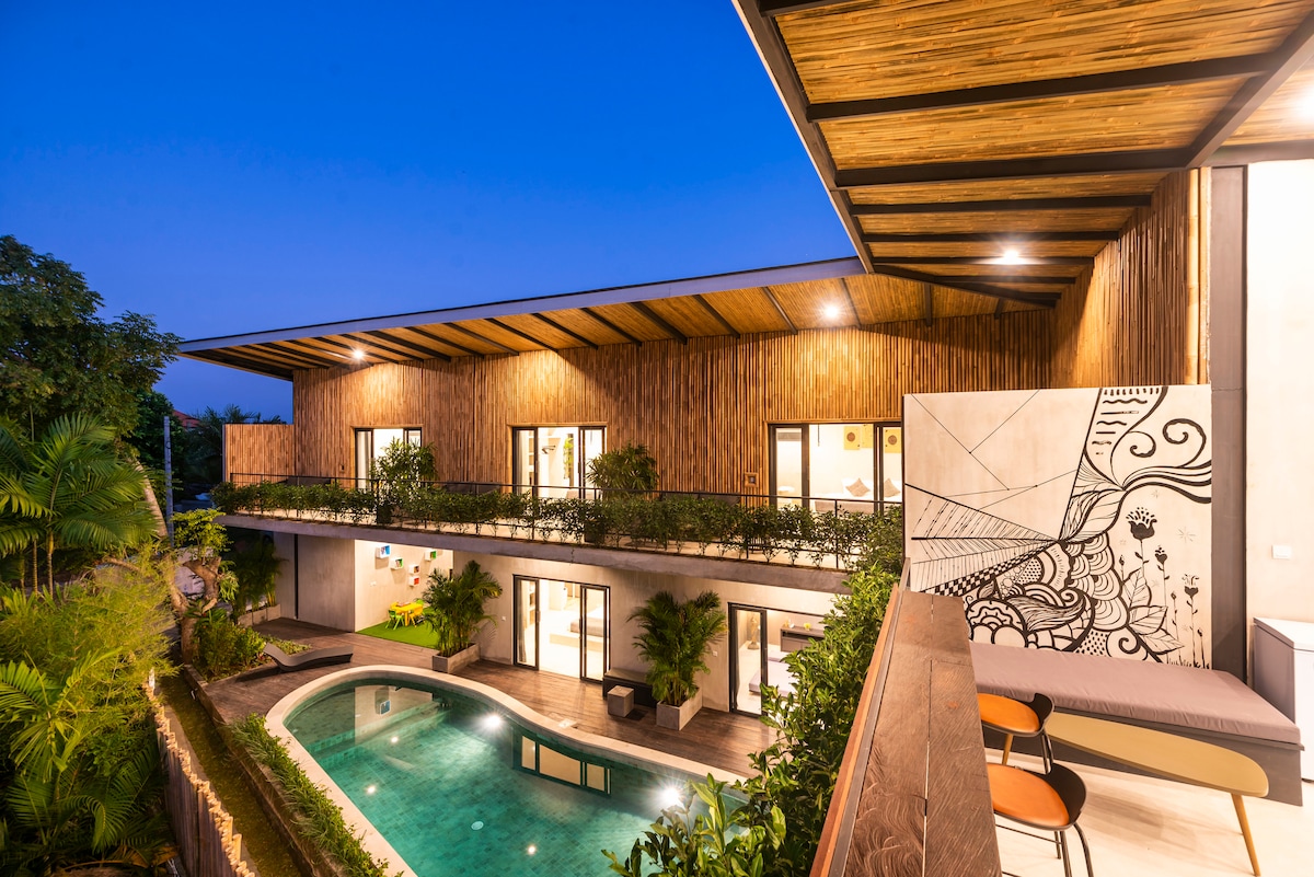 A modern building featuring wooden accents and expansive glass doors is situated alongside a tranquil swimming pool. Lush greenery surrounds the area, and the exterior is illuminated by evening light, enhancing the inviting ambiance of the space.