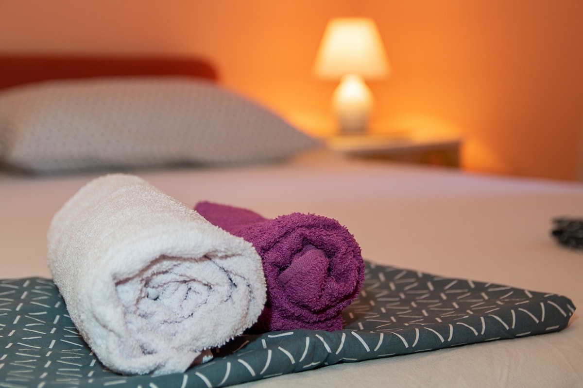 Neatly arranged bath towels are displayed on a bed with patterned bedding. A softly glowing lamp in the background provides warm lighting, while the vibrant wall color enhances the cozy ambiance of the space.