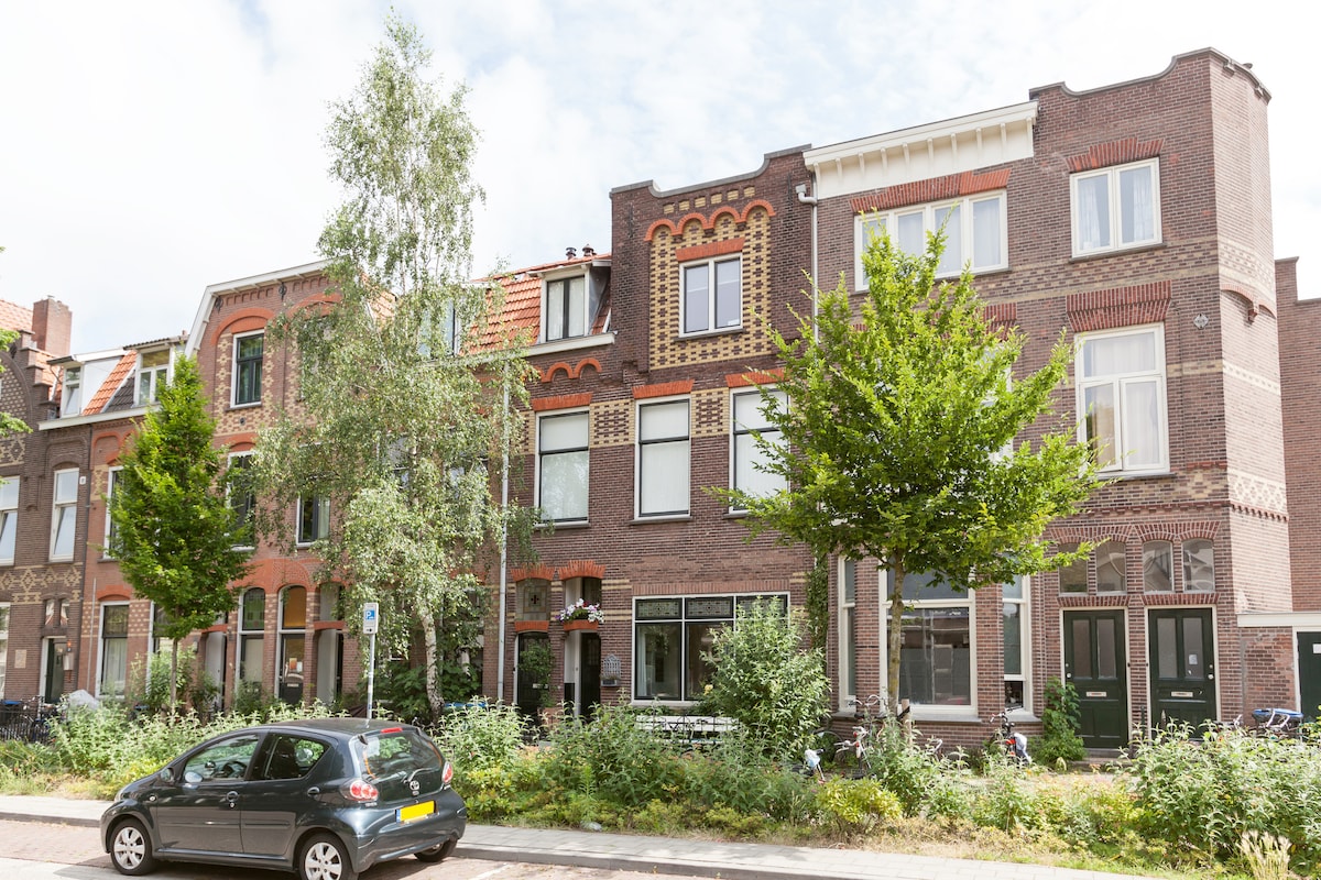 The image shows a charming street view of a three-story brick building, featuring intricate architectural details. Trees line the sidewalk, and green shrubs are present in the foreground. A parked car is visible at the street. The overall setting conveys a peaceful neighborhood atmosphere.