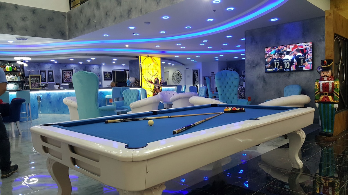 A spacious sports bar area features a blue pool table with cues resting on its edge. Neon lighting creates a vibrant atmosphere, highlighting modern seating and a bar counter in the background. A large screen displays a sports game, adding to the lively ambiance.