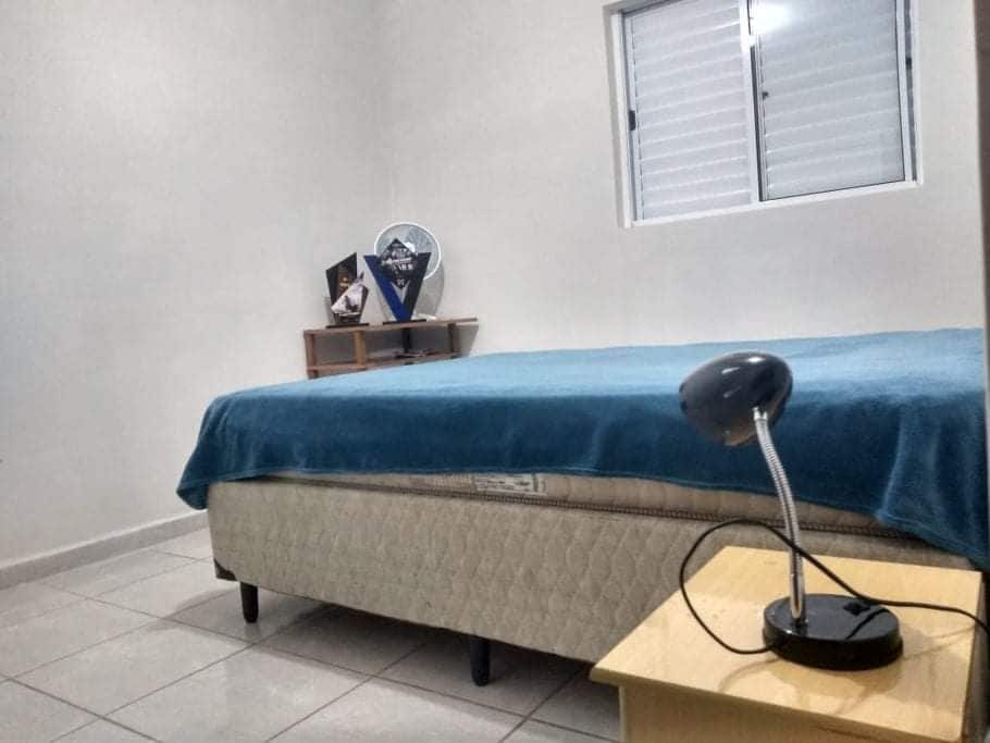 A well-furnished bedroom is featured, showcasing a spacious bed covered with a blue blanket. A bedside lamp is positioned on a small table next to the bed. A window brings in natural light, with a fan and decorative items visible on a shelf.