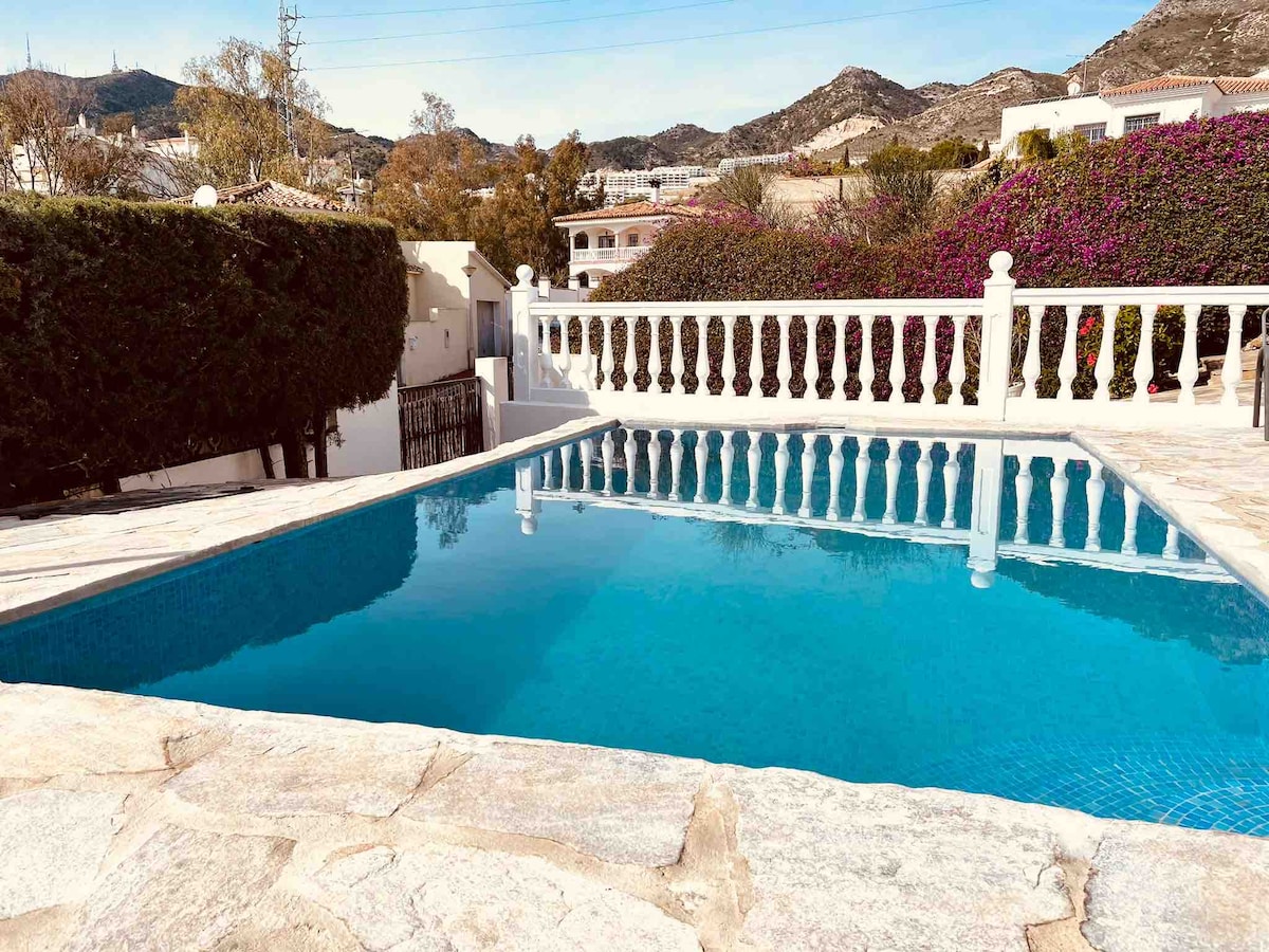 A well-maintained pool surrounded by a stone deck is visible, reflecting the bright blue sky. Lush greenery lines the pool area, enhancing the serene environment. The backdrop features hills, adding to the peaceful atmosphere of the shared outdoor space.