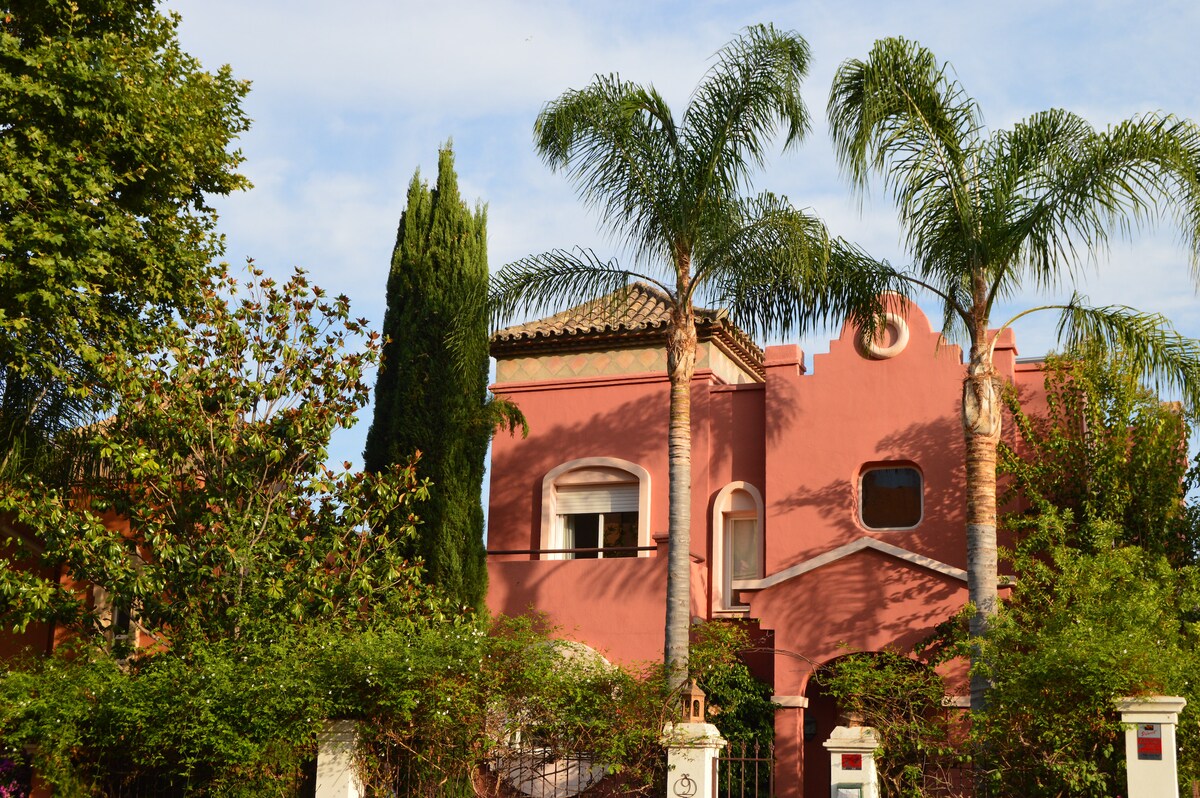 The villa features a charming pink exterior complemented by lush greenery and tall palm trees. Architectural details include a decorative roof and arched windows, providing an inviting appearance. The surrounding garden is well-maintained, enhancing the villa's appealing presence in its natural setting.