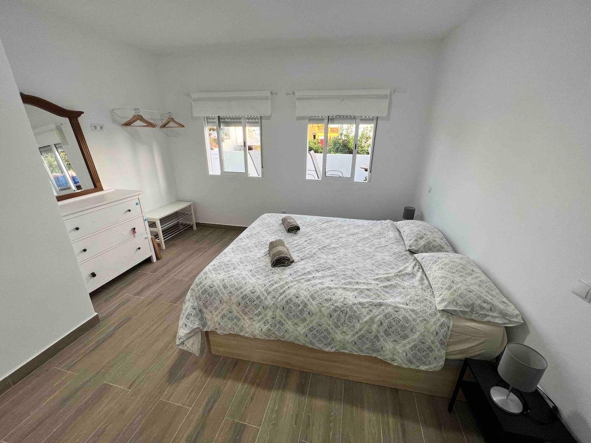A well-lit bedroom features a spacious bed adorned with light-colored linens. Two windows allow natural light to brighten the room, while a dresser and a small seating area are positioned against the wall. A mirror hangs above the dresser, complementing the minimalist decor.