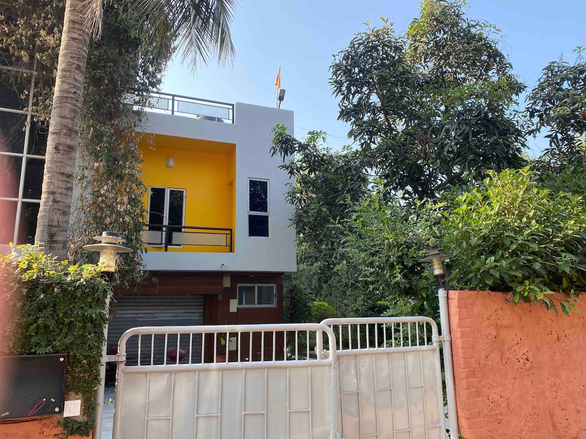 A contemporary house is surrounded by lush greenery, with a bright yellow facade visible through the white gate. Tall trees provide shade, creating a serene atmosphere. The entrance features a simple yet modern design, blending harmoniously with the natural elements in the environment.