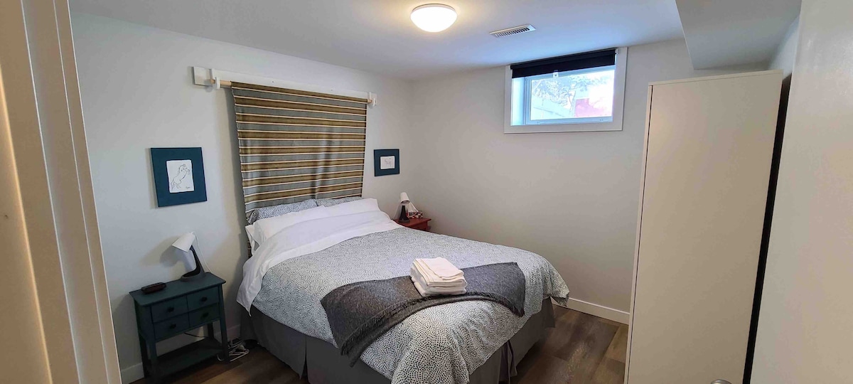 A cozy bedroom features a spacious bed dressed in soft linens. Natural light enters through a small window, with a decorative cover allowing for privacy. A bedside lamp and framed artwork add functional and visual interest, while a closet is positioned nearby.
