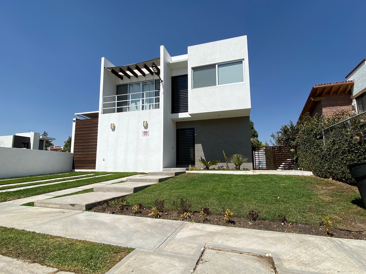 The modern house is showcased with a clean white facade and large windows. A well-kept lawn features a pathway leading to the entrance. The architectural design incorporates elements such as horizontal wood accents and clear landscaping, offering a welcoming first impression.
