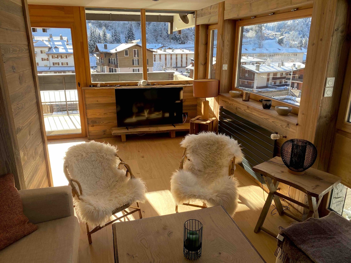 A cozy living area is created with wooden walls and large windows allowing natural light. Two comfortable armchairs with soft textiles are placed near a small table. A television is mounted on the wall, enhancing the inviting atmosphere of the space.