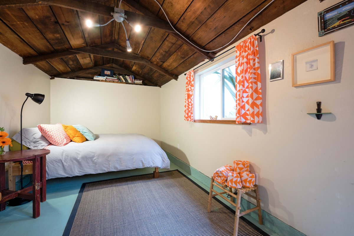 A cozy room is presented with a rustic wooden ceiling. A spacious bed is adorned with colorful pillows. A small table beside the bed features a lamp, while a patterned chair adds a pop of color. Natural light streams through the window, creating a bright atmosphere.