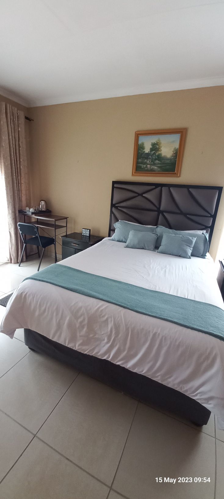 A well-appointed sleeping area features a spacious bed with a light teal throw, complemented by two pillows. A small desk and chair are positioned nearby. Natural light enters through a window, partially draped with cream curtains, against a neutral wall with a landscape painting.