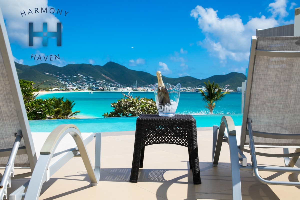 Two lounge chairs frame a stunning view of the Caribbean Sea, with a small side table holding a bucket of ice and champagne. The background features lush hills and a vibrant blue sky, creating a serene outdoor setting.