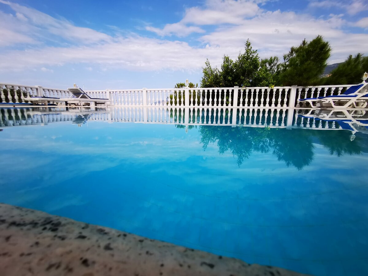 A tranquil pool is depicted, surrounded by a white railing. Sun loungers are positioned nearby, providing a space for relaxation. The clear blue water reflects the sky, creating a serene atmosphere. Lush greenery can be seen in the distance, enhancing the natural setting.
