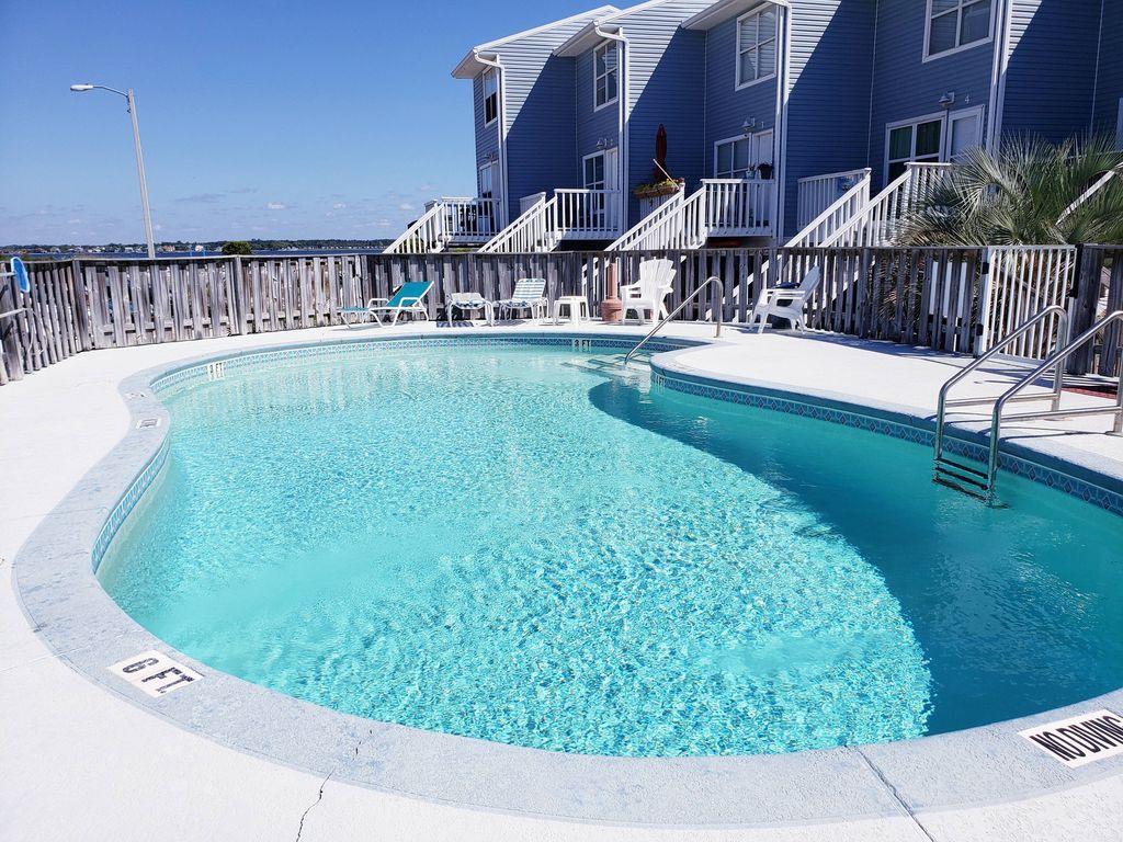 A swimming pool is set in a well-maintained area, surrounded by a wooden deck and lounge chairs. The water is clear and inviting, reflecting the blue sky above. The townhouse structure is visible in the background, featuring multiple units with balconies.