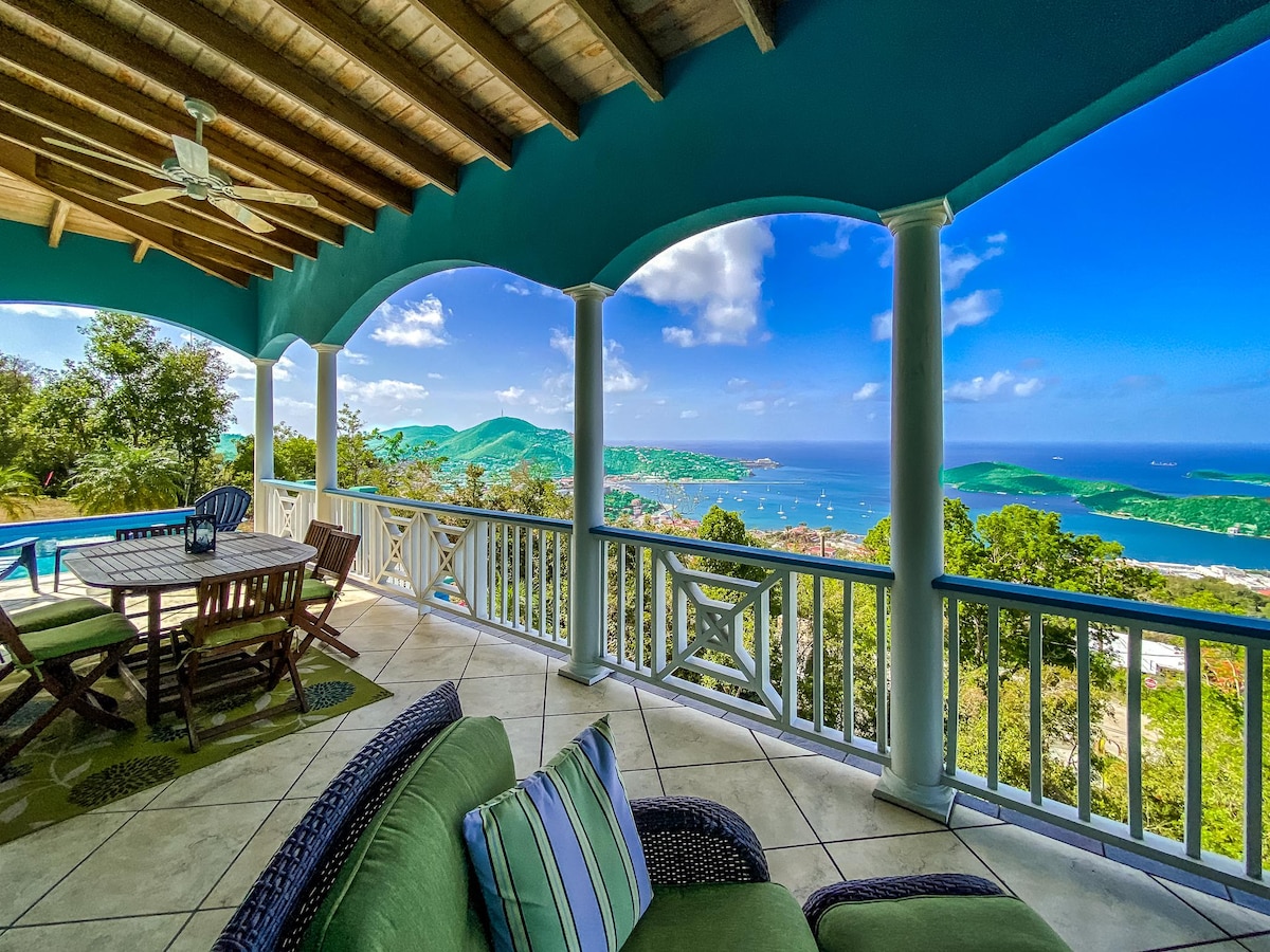 A covered outdoor seating area is furnished with a large dining table and comfortable lounge chairs. The space offers sweeping views of Charlotte Amalie Harbor, surrounded by lush greenery and rolling hills. The blue sky and distant islands create a serene backdrop.