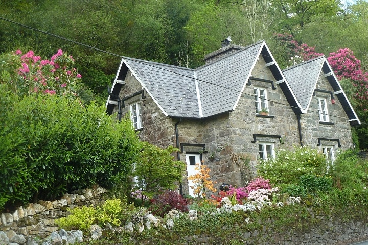 A charming stone cottage is surrounded by lush greenery and colorful flowering plants. The unique roofline features multiple peaks, while the entrance is framed by neatly arranged shrubs. The natural landscape offers a serene backdrop, enhancing the peaceful setting of the cottage.