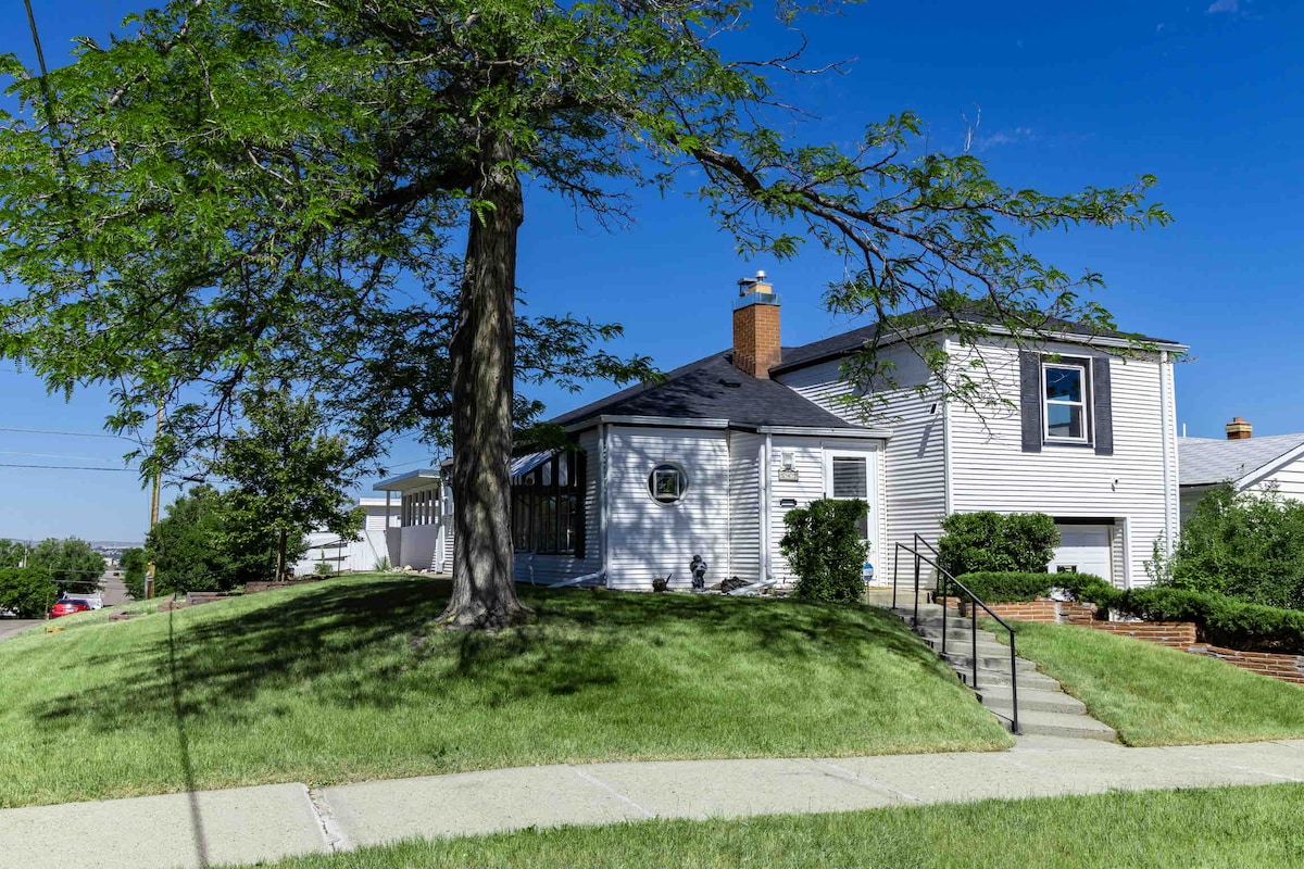 The exterior of the home is shown, featuring a mid-century design with a sloped roof and multiple windows. A large tree stands in front, providing shade. The landscape is well-maintained with green grass and a gentle slope leading to a set of stairs.