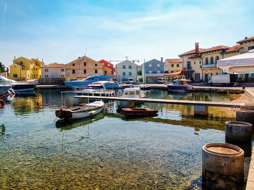 The image captures a serene harbor scene with several boats anchored in clear waters. Colorful buildings line the waterfront, reflecting a blend of traditional and contemporary architecture. The gentle ripples on the water and the blue sky above enhance the tranquil atmosphere.