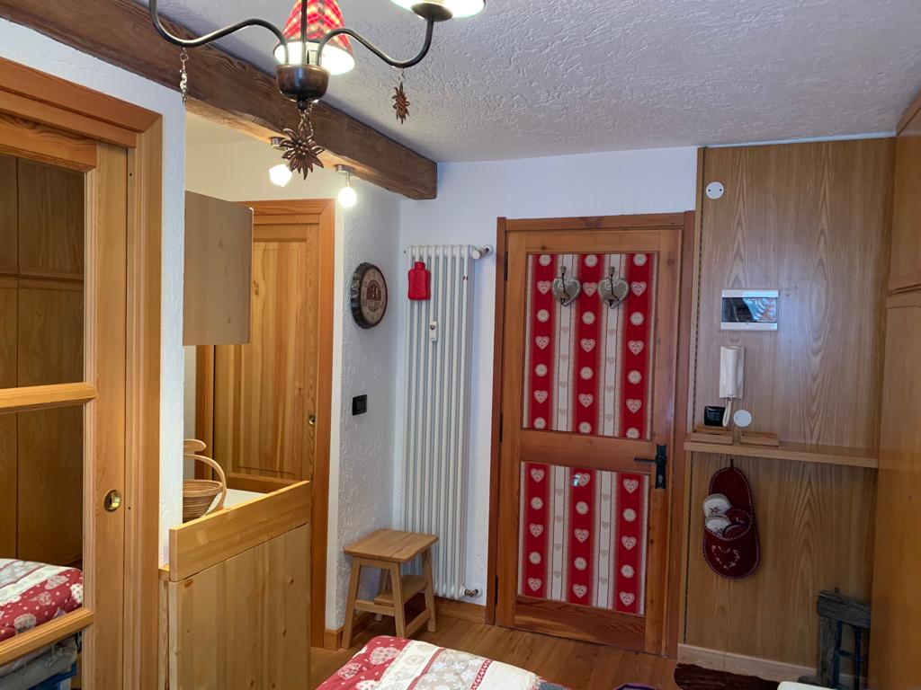 The cozy interior features wooden accents and warm tones. A traditional red door adorned with hearts complements the rustic style. A small stool is positioned near a wooden partition, with a radiator visible. Decorative elements include a wall hanging and holiday ornaments.